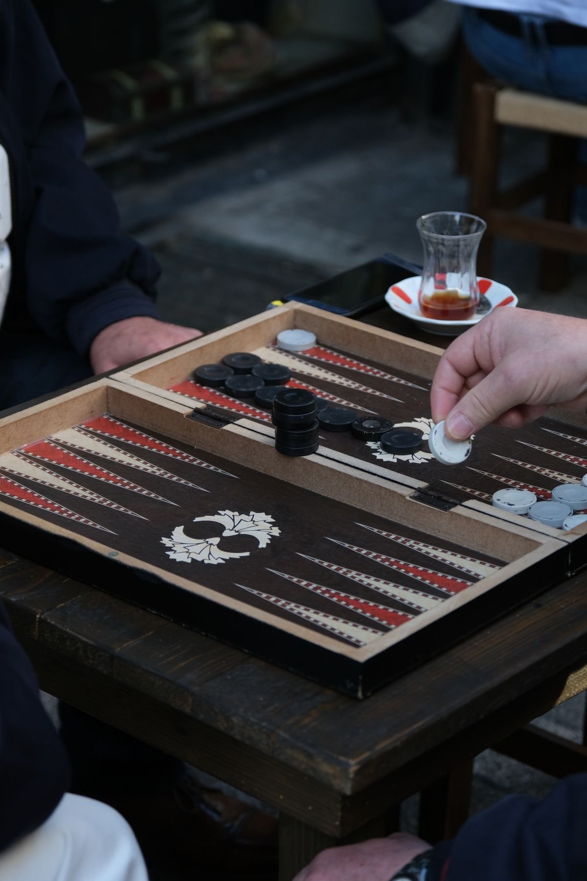 A hand moving a checker on a full backgammon board at an outdoor cafe table, with a glass of tea beside it